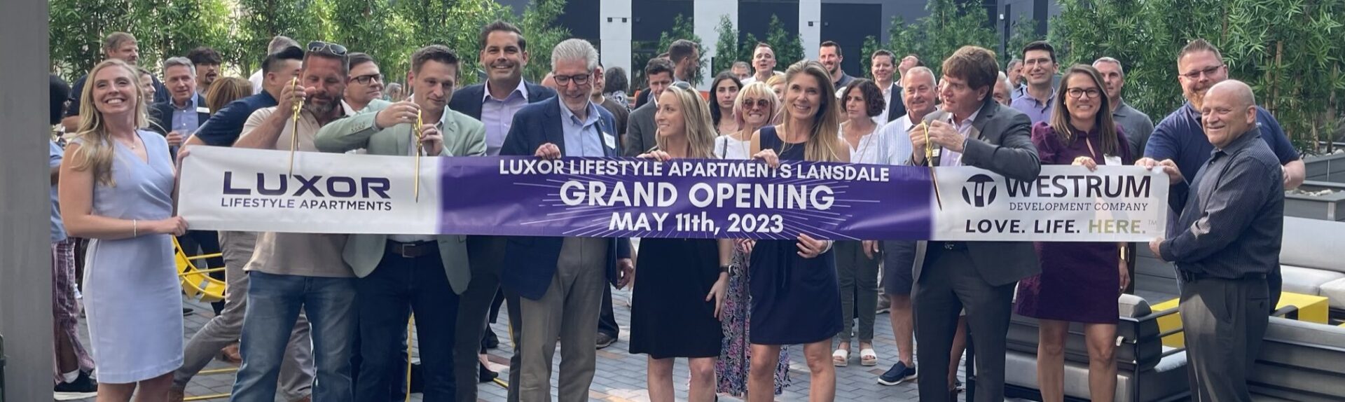 Westrum Corporate A group of people smile and pose with a "Grand Opening" banner for Luxor Lifestyle Apartments, May 11th, 2023.