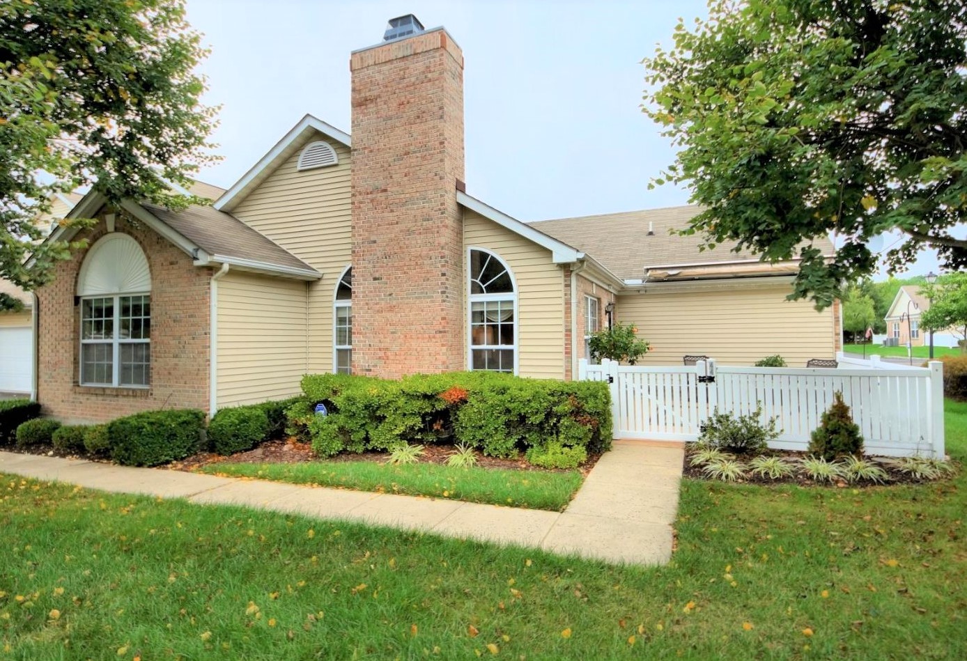 Westrum Corporate Charming Lawrence Township single-story with brick chimney, tan siding, white fence, lush shrubs, mature trees, and a well-kept lawn.