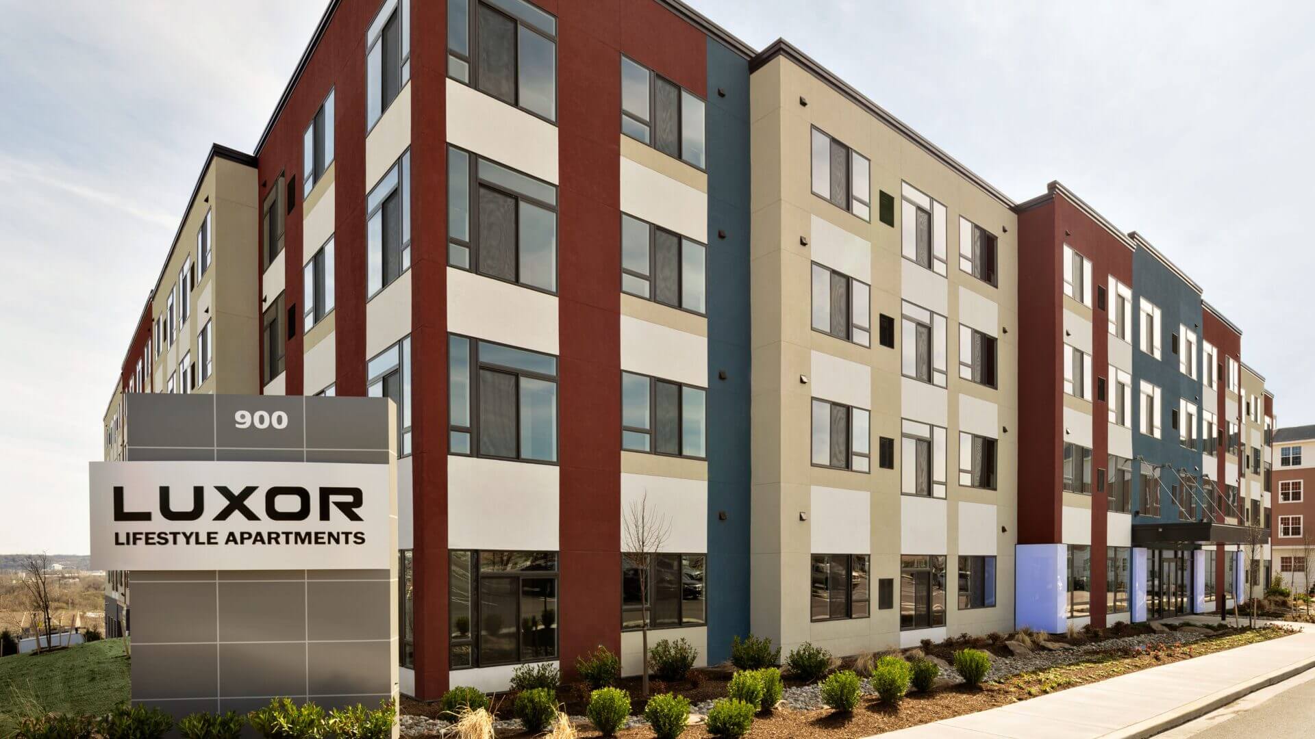 Westrum Corporate Modern four-story building with red, beige, and white accents; Luxor Lifestyle Apartments sign stands prominently in front.