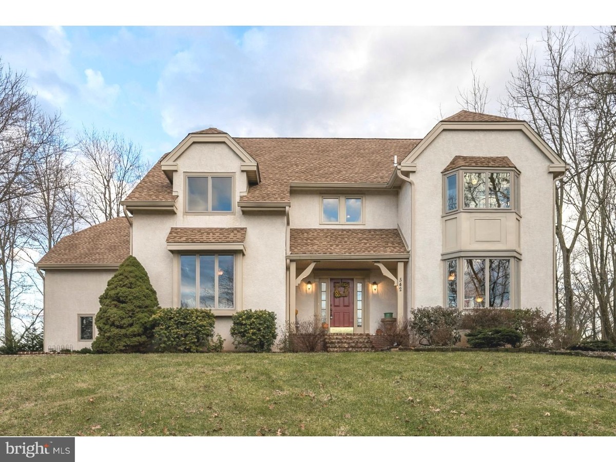 Westrum Corporate Two-story beige house in Skippack Township with large windows, brown roof, red door, shrubs, and trees on a grassy lawn.