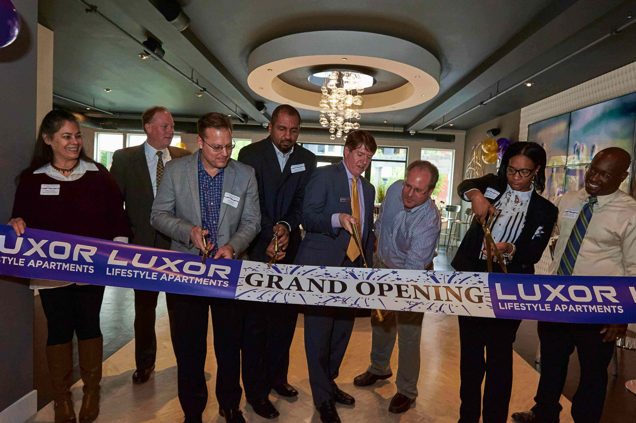 Westrum Corporate Eight people cut a "Grand Opening" ribbon inside a modern apartment building, celebrating Luxor Lifestyle Apartments.