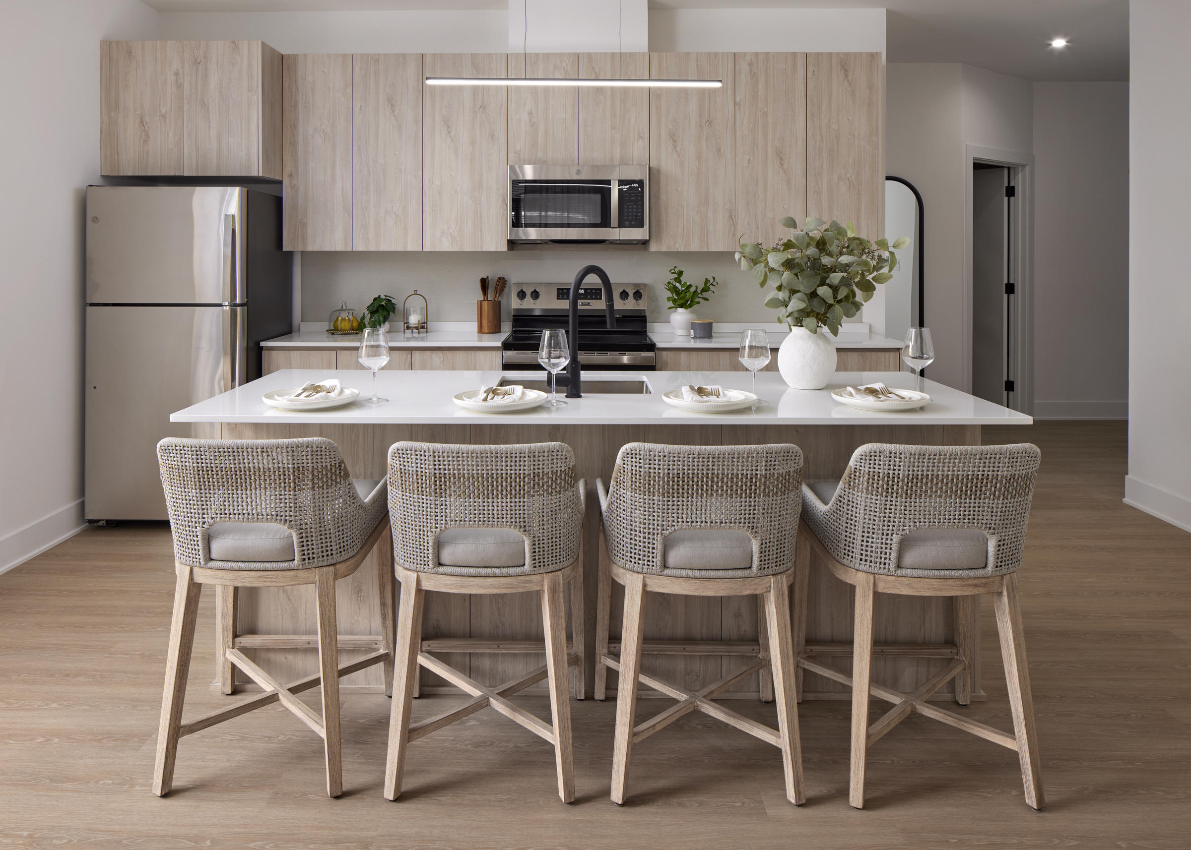 Westrum Corporate Modern kitchen with wood cabinets, four stools at an island, stainless steel appliances, and a vase with greenery on the counter.