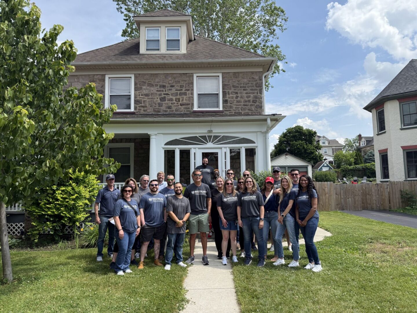Westrum Corporate A group of people stand smiling on a sidewalk in front of a two-story stone house on a sunny day.