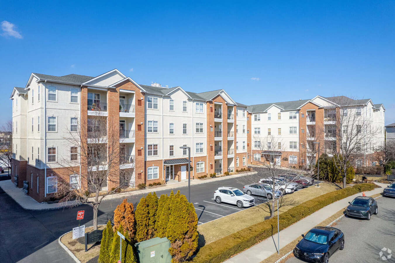 Westrum Corporate Regency at Packer Park: Four-story brick and beige apartment, parking lot, cars, and trimmed bushes under a clear blue Philadelphia sky.