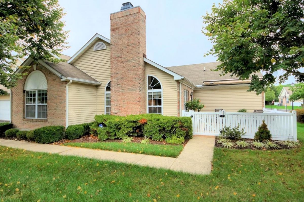 Westrum Corporate Charming Lawrence Township single-story with brick chimney, tan siding, white fence, lush shrubs, mature trees, and a well-kept lawn.