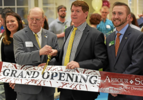 Westrum Corporate Four people smile as one cuts a large "Grand Opening" ribbon at an event, with a crowd and balloons in the background.