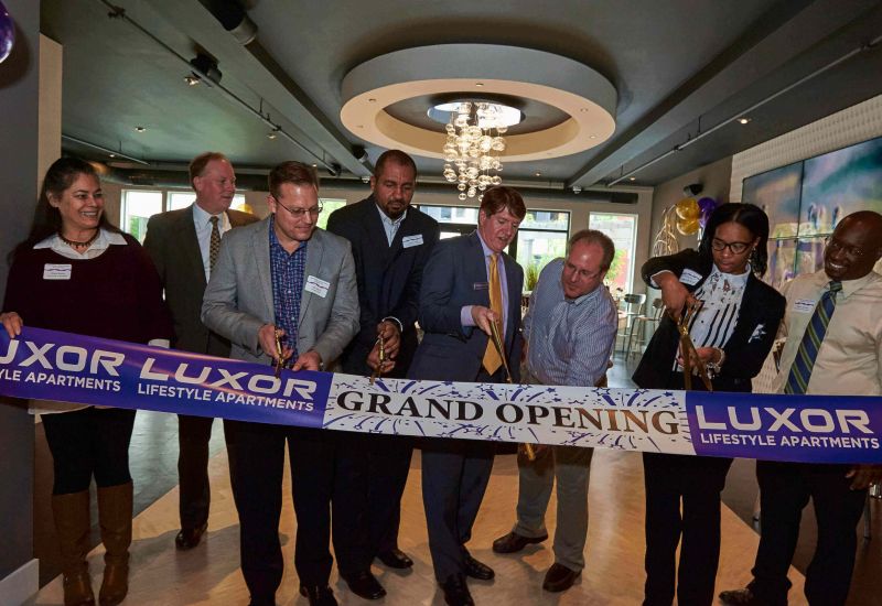 Westrum Corporate Eight people cut a "Grand Opening" ribbon inside a modern apartment building, celebrating Luxor Lifestyle Apartments.