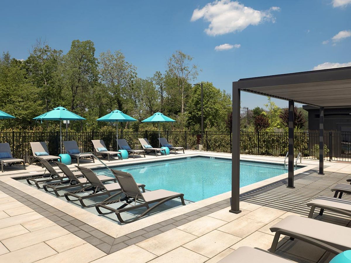 Westrum Corporate Relax at the Foundry Flats outdoor pool, surrounded by lounge chairs, blue umbrellas, lush trees, and a sunny sky.