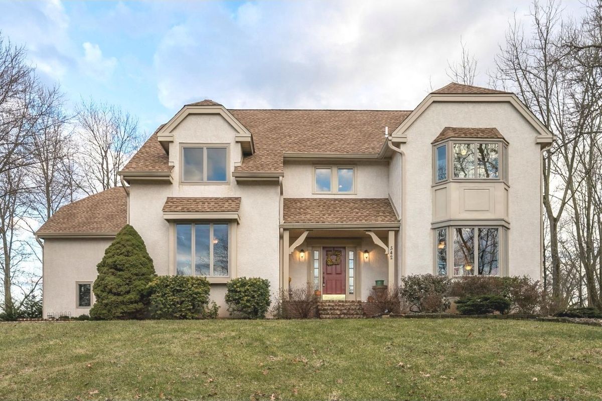Westrum Corporate Two-story beige house in Skippack Township with large windows, brown roof, red door, shrubs, and trees on a grassy lawn.