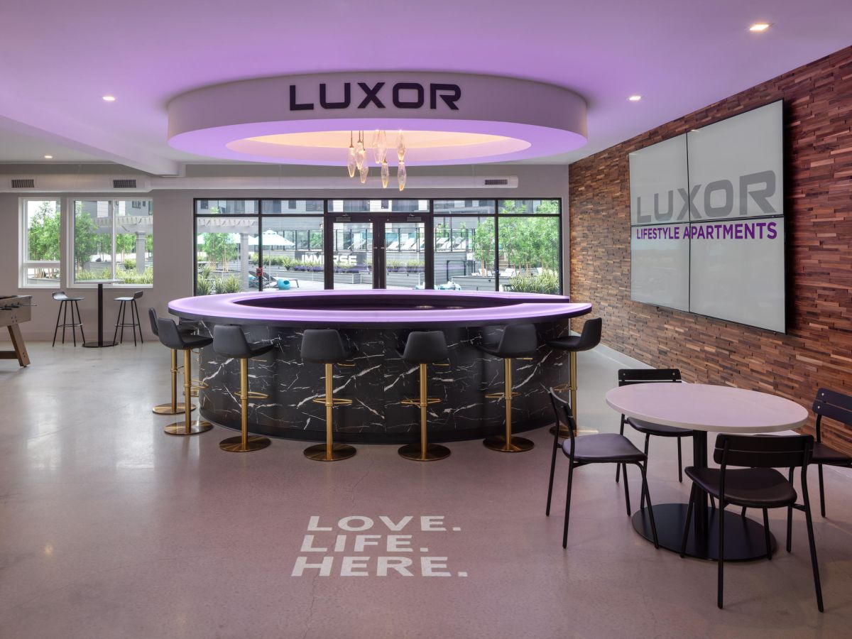 Westrum Corporate Modern lounge at Luxor Montgomeryville: circular bar, black chairs, “LUXOR” sign, and “LOVE. LIFE. HERE.” on the floor with wood accents.