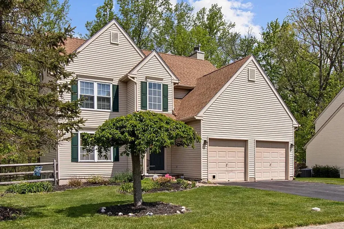Westrum Corporate Two-story beige house in Buckingham Square with green shutters, double garage, manicured lawn, and a small tree in the sunny front yard.