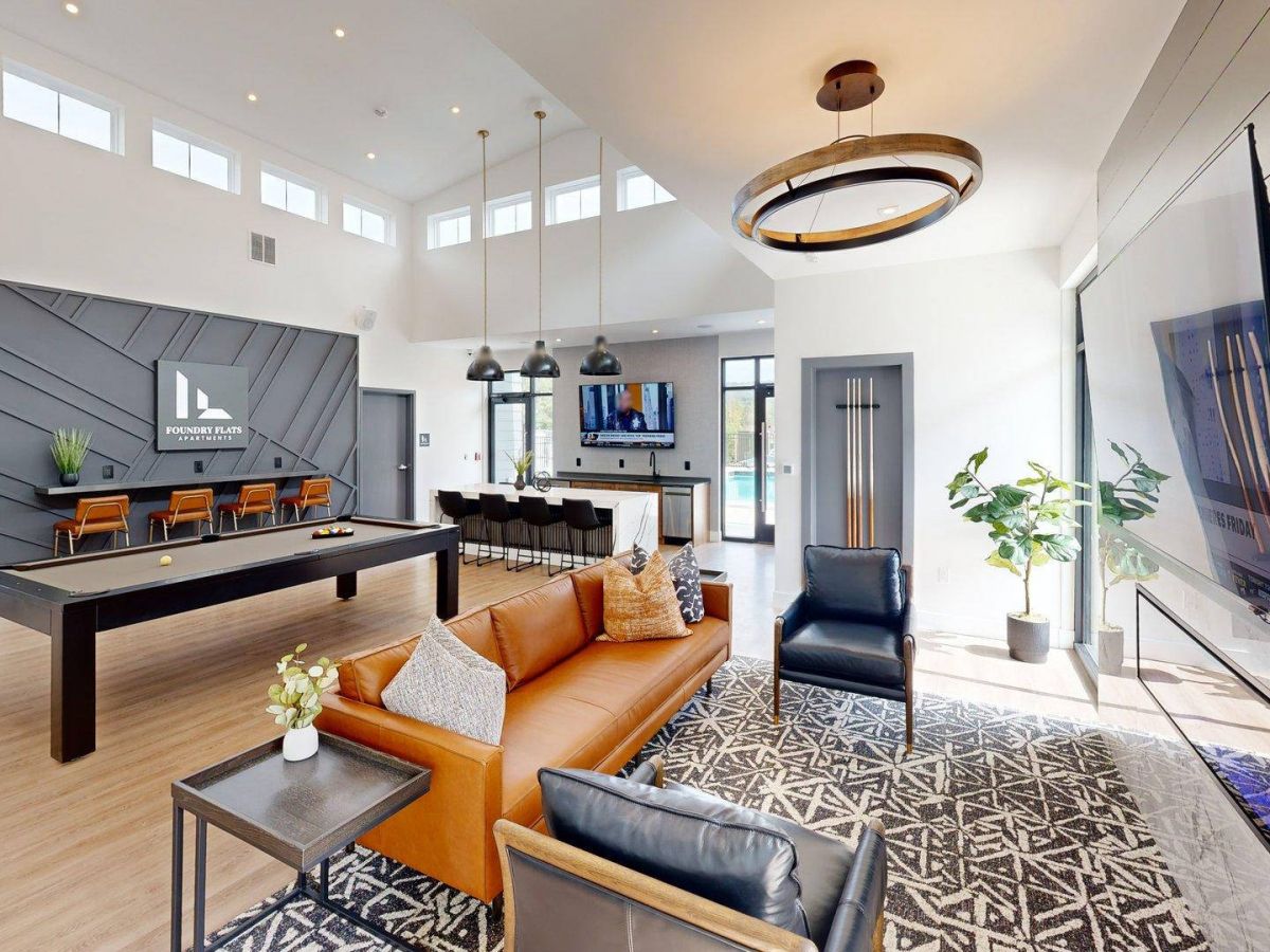 Westrum Corporate Modern lounge with pool table, brown sofa, black armchair, kitchen area, large windows, plants, and wall-mounted TV.
