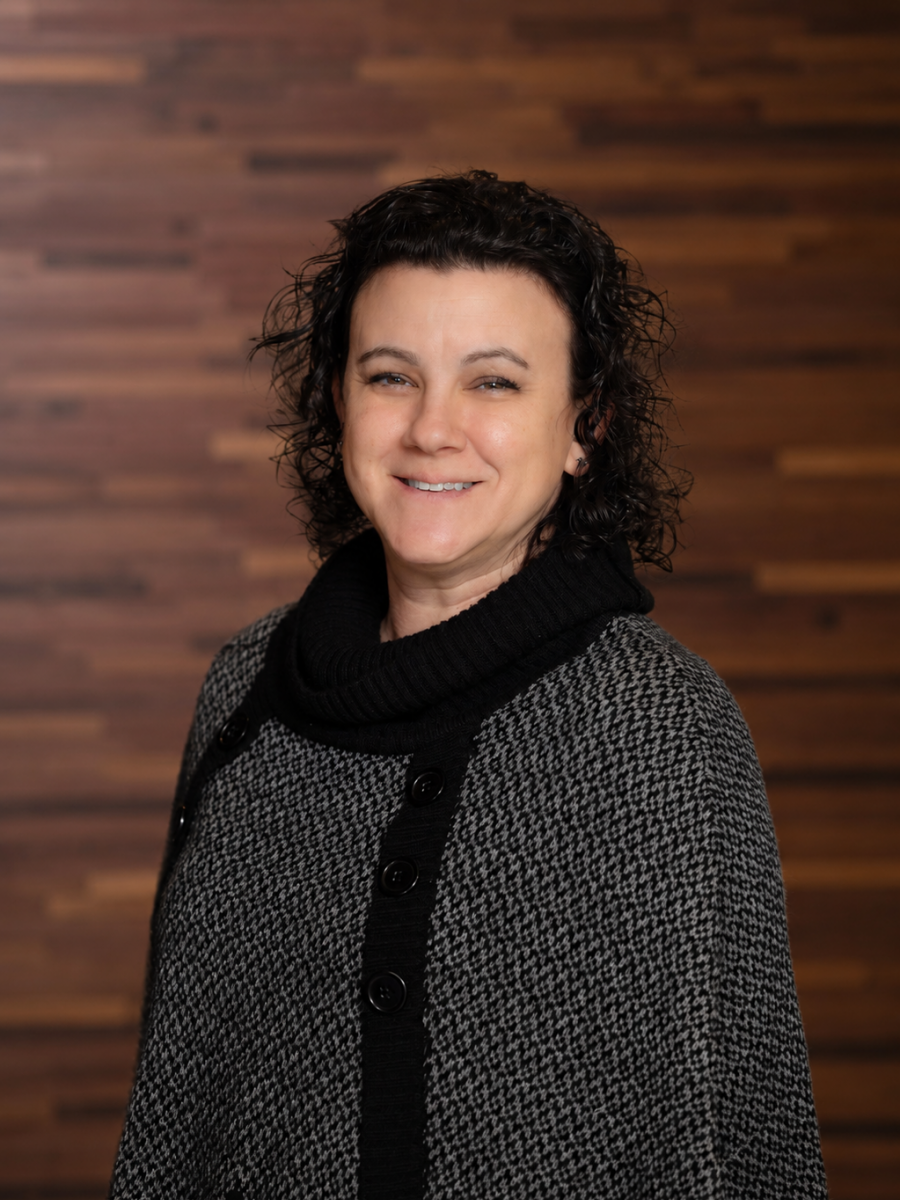 Westrum Corporate Christine Yordy, a woman with curly dark hair, stands smiling in a gray and black sweater before a wooden wall background.