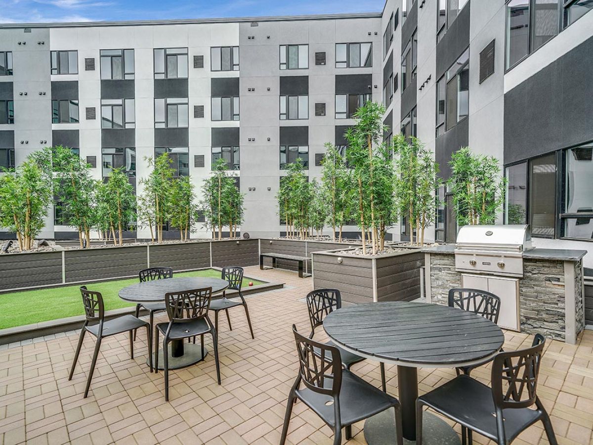 Westrum Corporate Modern outdoor patio at Luxor West Chester with round tables, grill, planters with trees, and nearby apartment buildings.