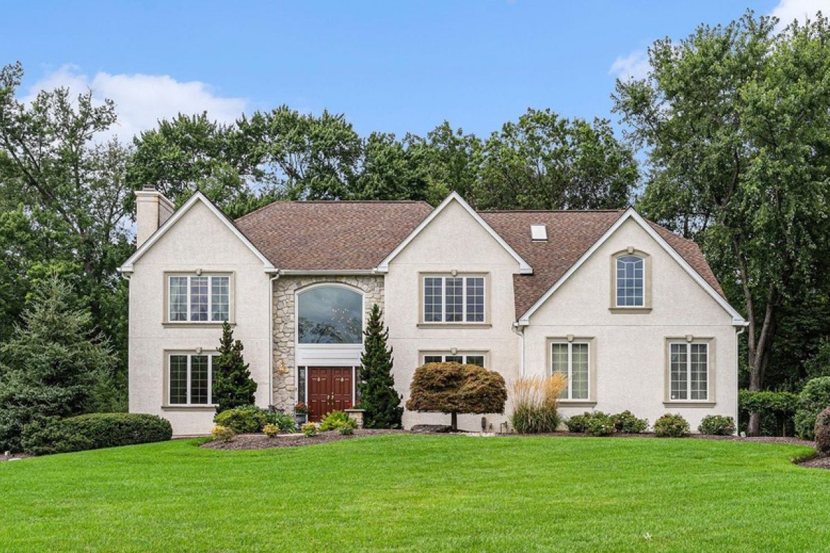 Westrum Corporate Two-story beige house with a brown roof, large windows, double front doors, and landscaped green lawn with bushes and trees.