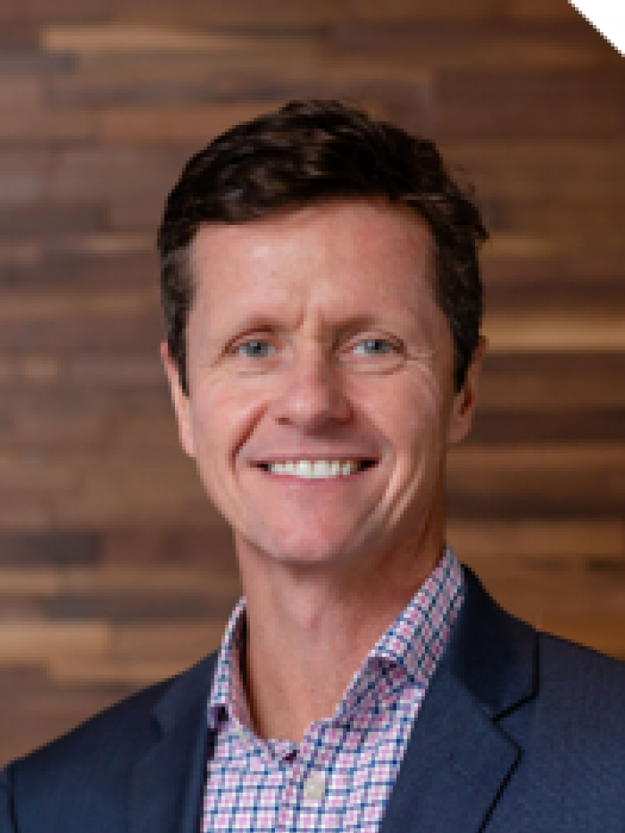 Westrum Corporate Michael Maier, in a blue suit and checked shirt, smiles warmly while standing before a wooden wall background.