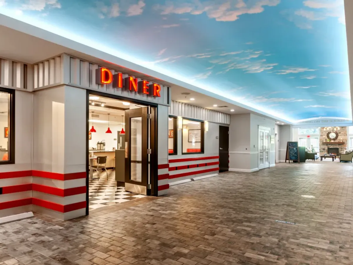 Westrum Corporate A retro diner with red accents and neon sign brightens Arbour Square senior living’s hallway, beneath a sky-themed ceiling.