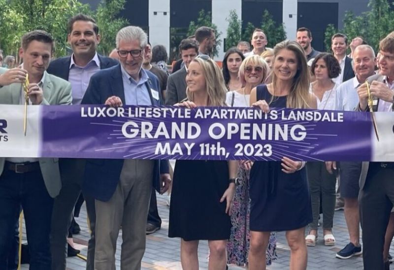 Westrum Corporate A group of people smile and pose with a "Grand Opening" banner for Luxor Lifestyle Apartments, May 11th, 2023.