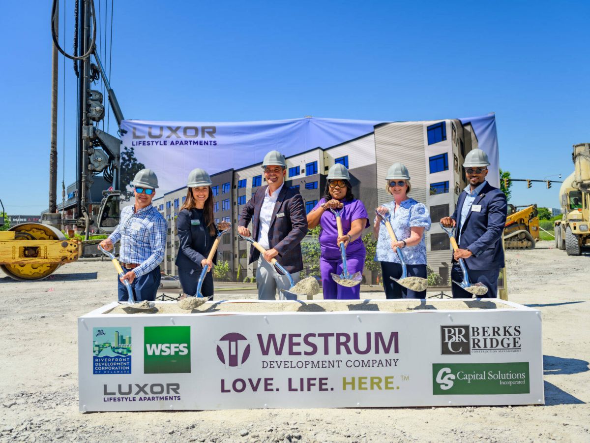 Westrum Corporate Six people in hard hats pose with shovels at a groundbreaking ceremony for Luxor Lifestyle Apartments, with construction equipment behind them.