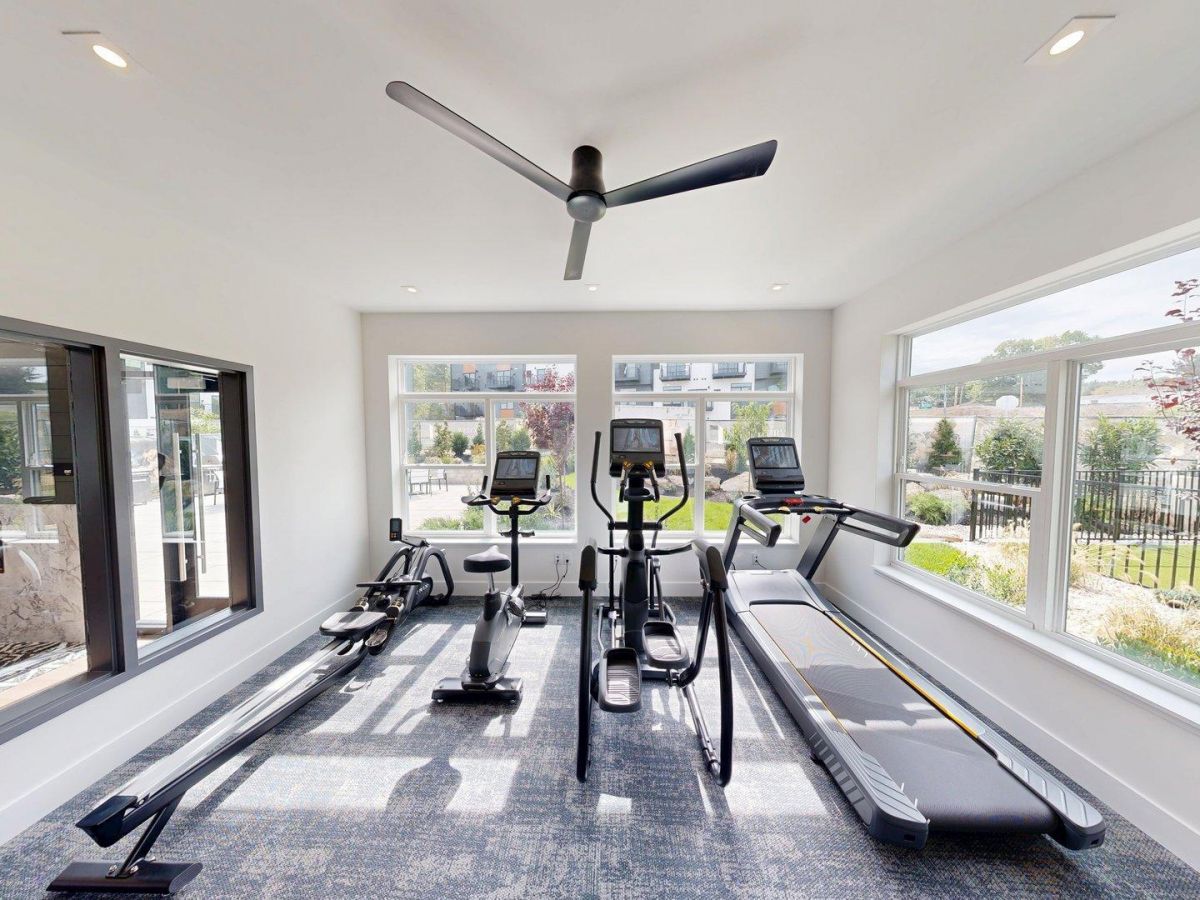 Westrum Corporate Foundry Flats small gym room with rowing machines, treadmills, large windows, ceiling fan, and views of greenery outside.