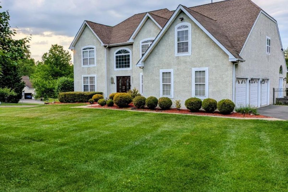 Westrum Corporate Two-story beige house with a brown roof, well-kept lawn, shrubs, and a three-car garage under a cloudy sky.
