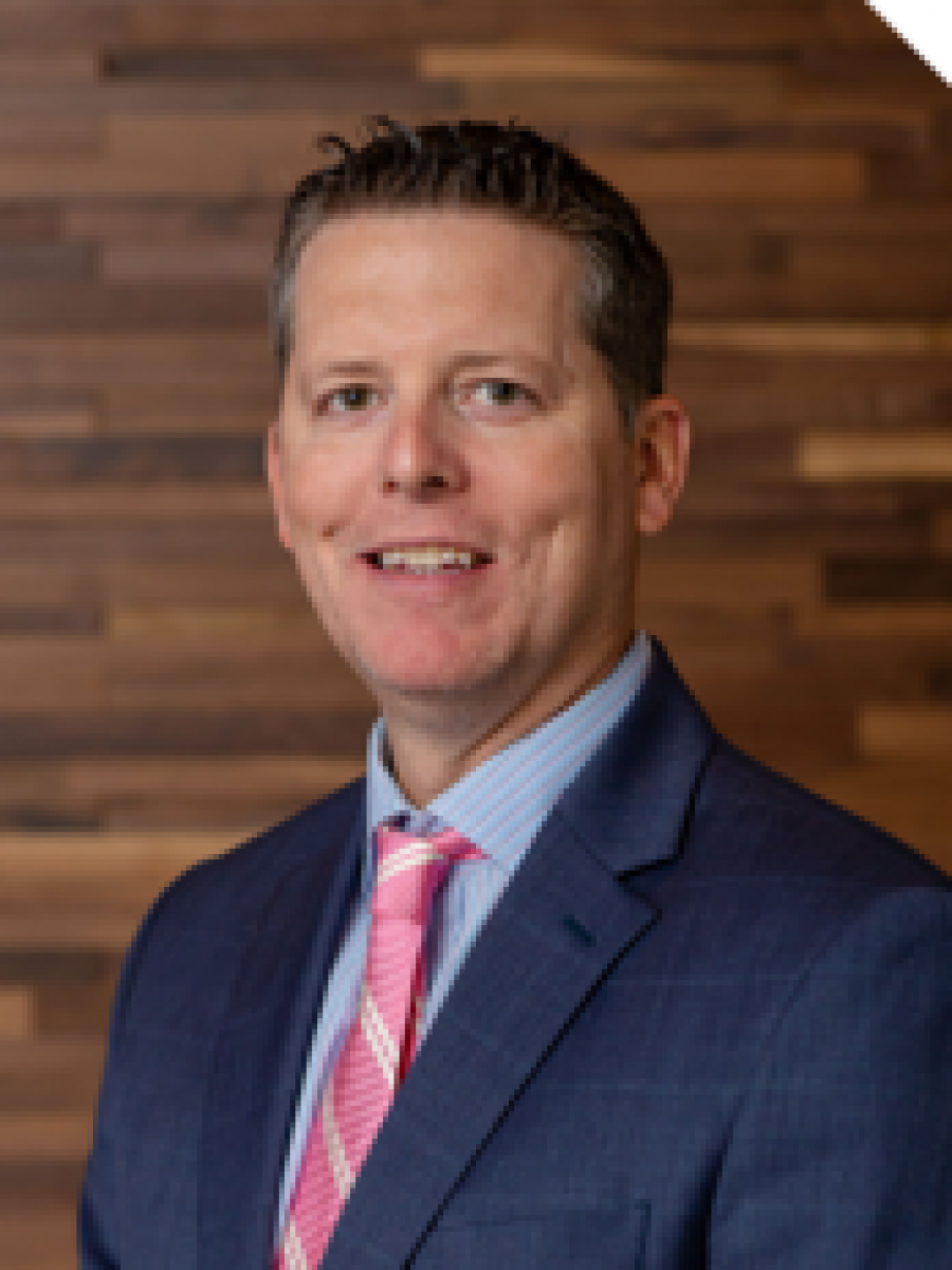 Westrum Corporate Brian McKenzie, in a blue suit and pink tie, stands smiling at the camera before a wooden wall.