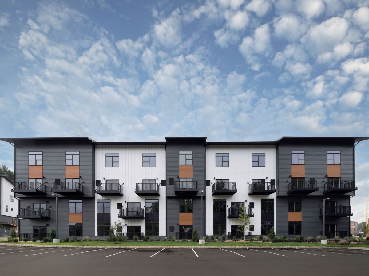 Modern three-story apartment building with balconies, parking spaces, and a partly cloudy sky above.