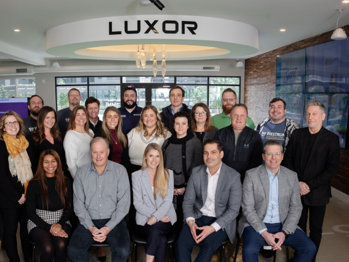 Westrum Corporate A group of twenty-one adults pose for a photo in an office lobby under a sign that says "LUXOR.