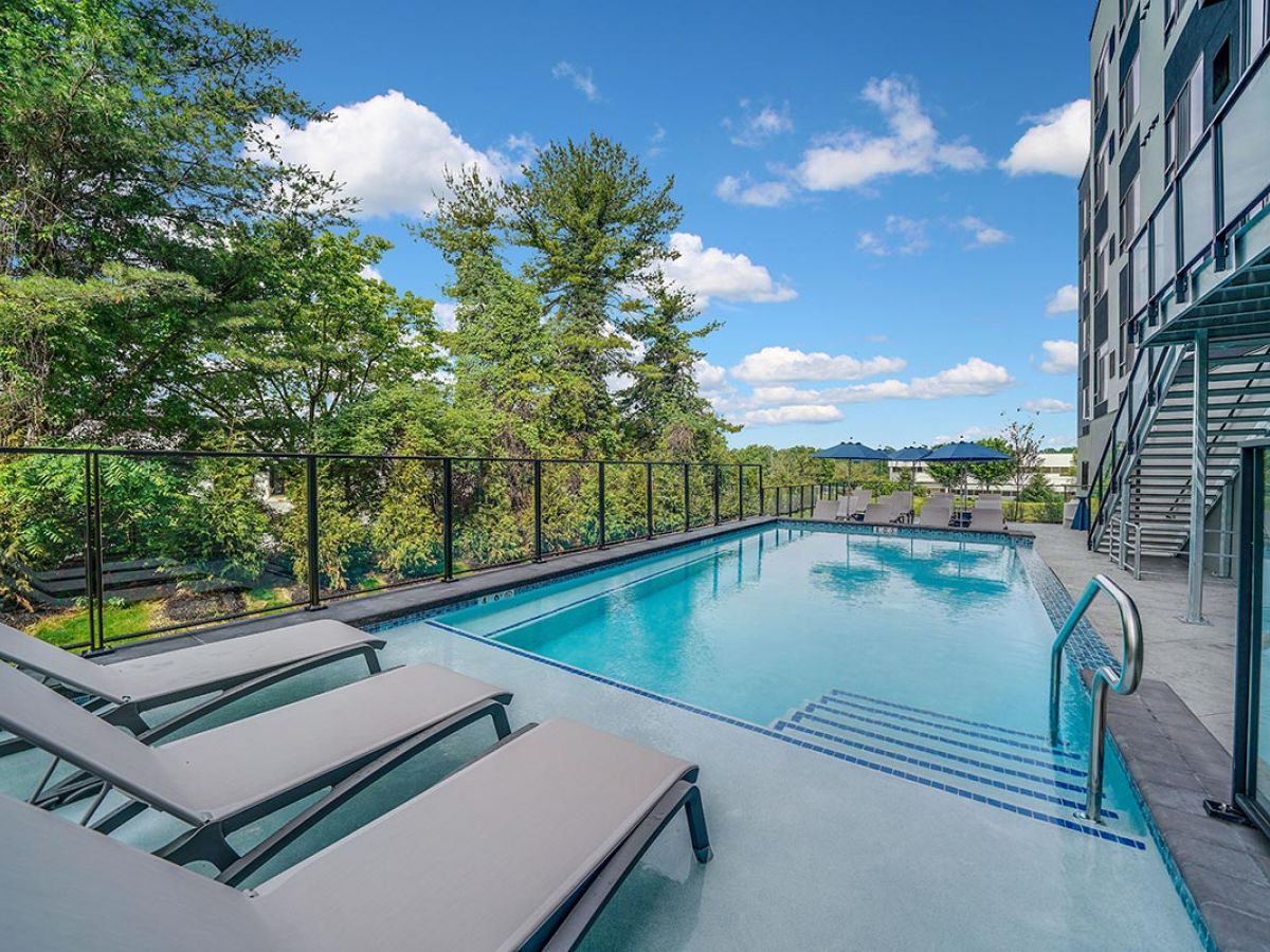 Westrum Corporate Luxor West Chester features an outdoor pool with lounge chairs, glass fence, trees, and a modern building under a blue sky.
