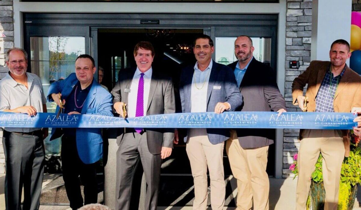 Westrum Corporate Eight people smile and hold a blue ribbon at a business ribbon-cutting ceremony in front of a building entrance.