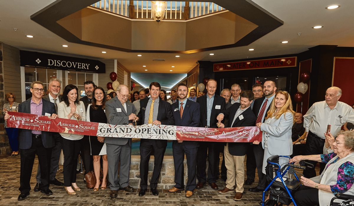 Westrum Corporate A group of people in business attire hold a ribbon that reads "Grand Opening" inside a building, with a crowd gathered.