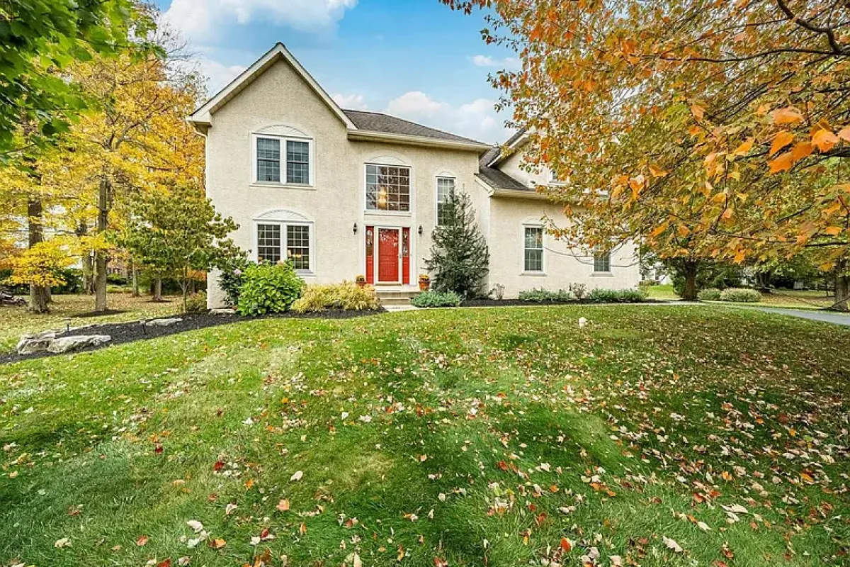 Westrum Corporate Two-story beige house in Upper Gwynedd Township with red doors, large windows, and a green lawn bordered by autumn’s orange-yellow trees.