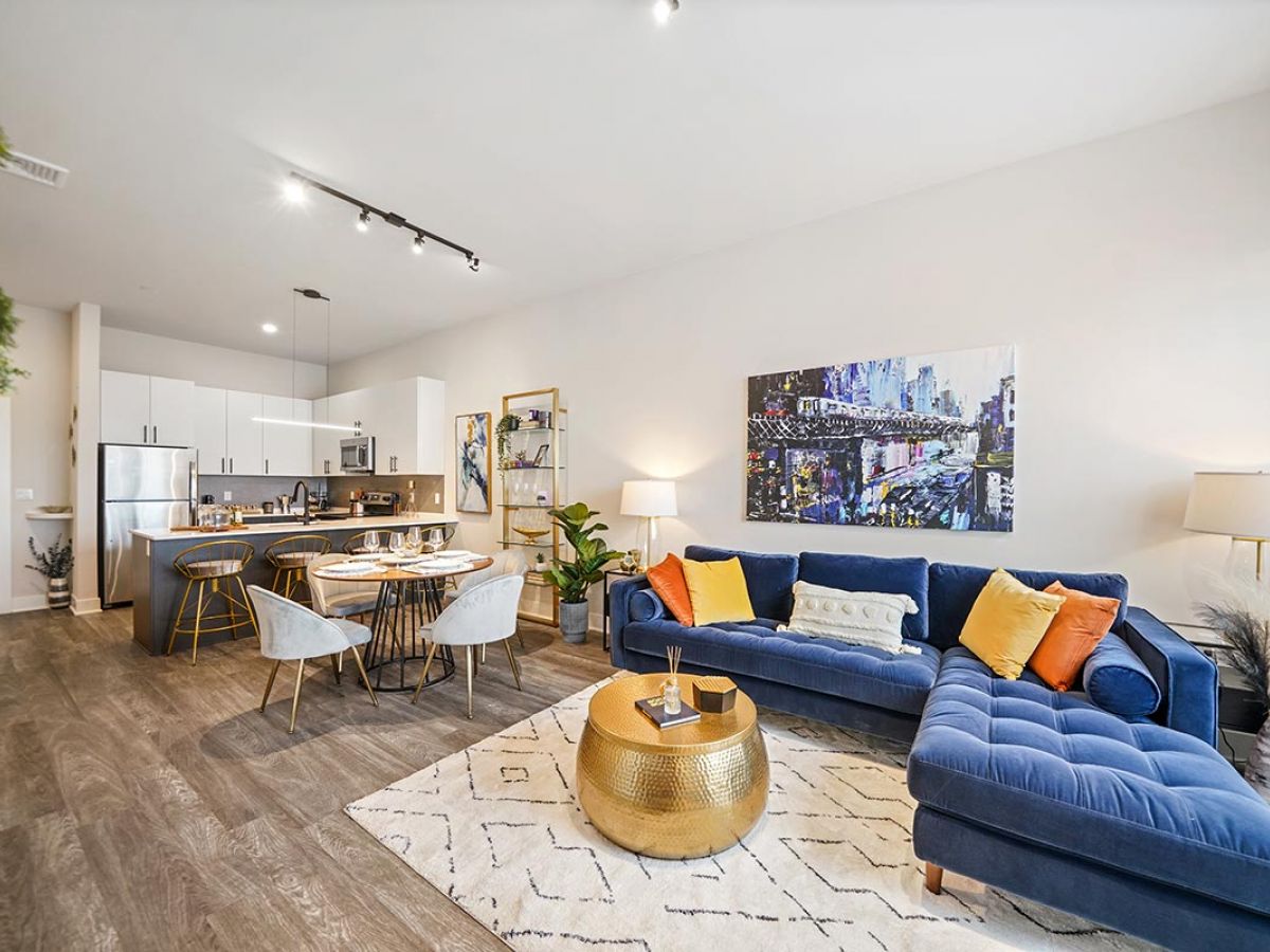 Westrum Corporate Modern open-plan living at Luxor Lansdale with blue sectional sofa, gold table, dining area, and cityscape artwork on the wall.