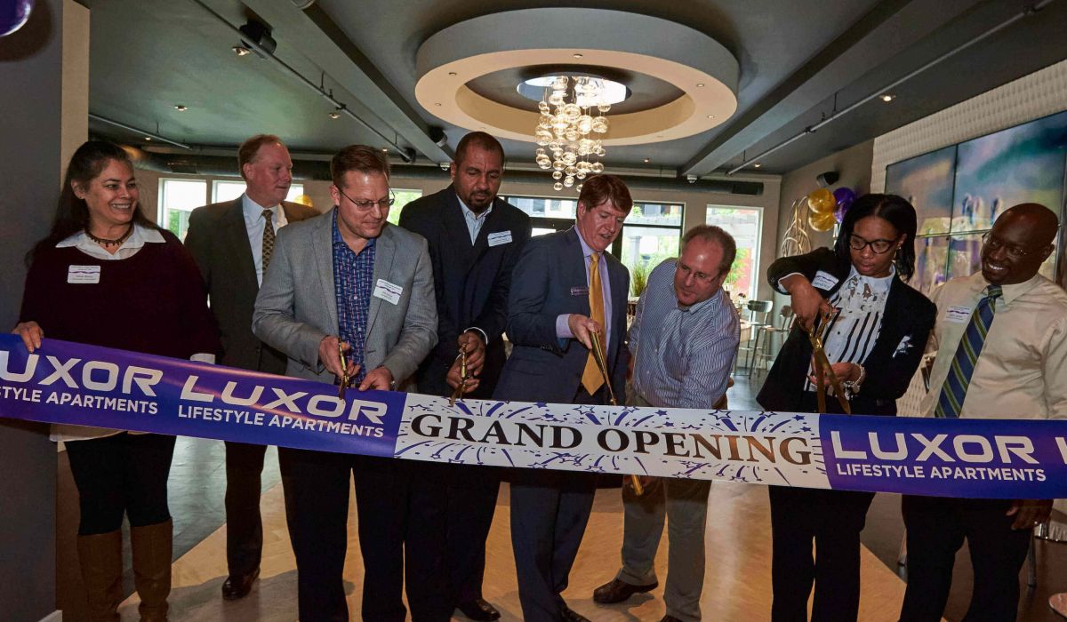 Westrum Corporate Eight people cut a "Grand Opening" ribbon inside a modern apartment building, celebrating Luxor Lifestyle Apartments.