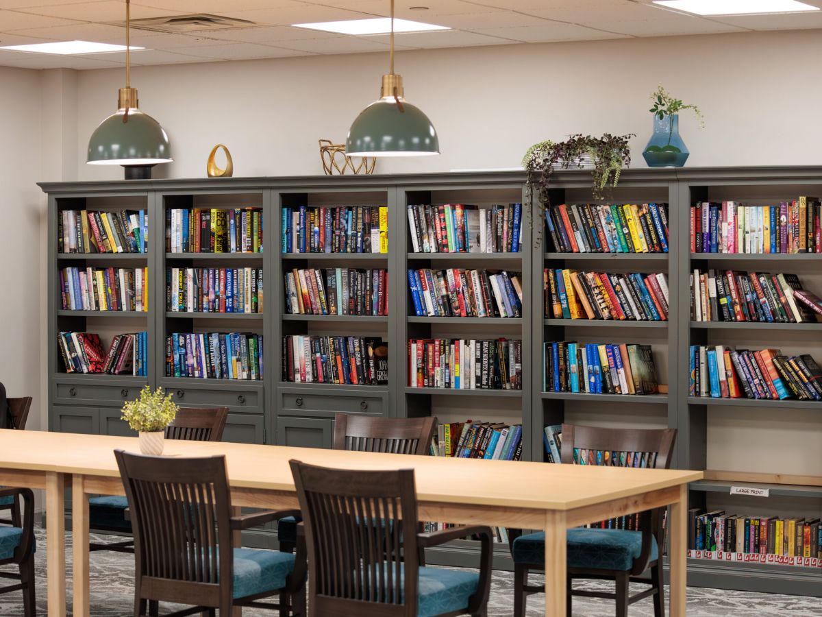 Westrum Corporate Bookshelves filled with colorful books behind a wooden table in Arbour Square’s well-lit senior living library room in Harleysville.