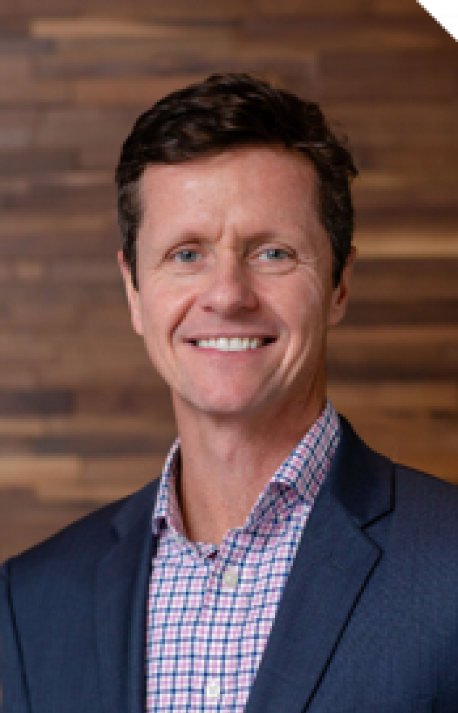 Westrum Corporate Michael Maier, in a blue suit and checked shirt, smiles warmly while standing before a wooden wall background.