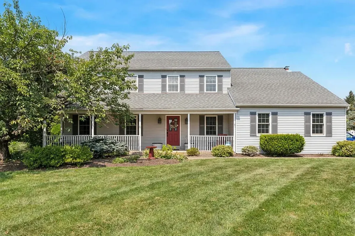 Westrum Corporate Two-story house with gray siding in Towamencin Township, red door, large lawn, green bushes, and bright blue sky in Montgomery County.