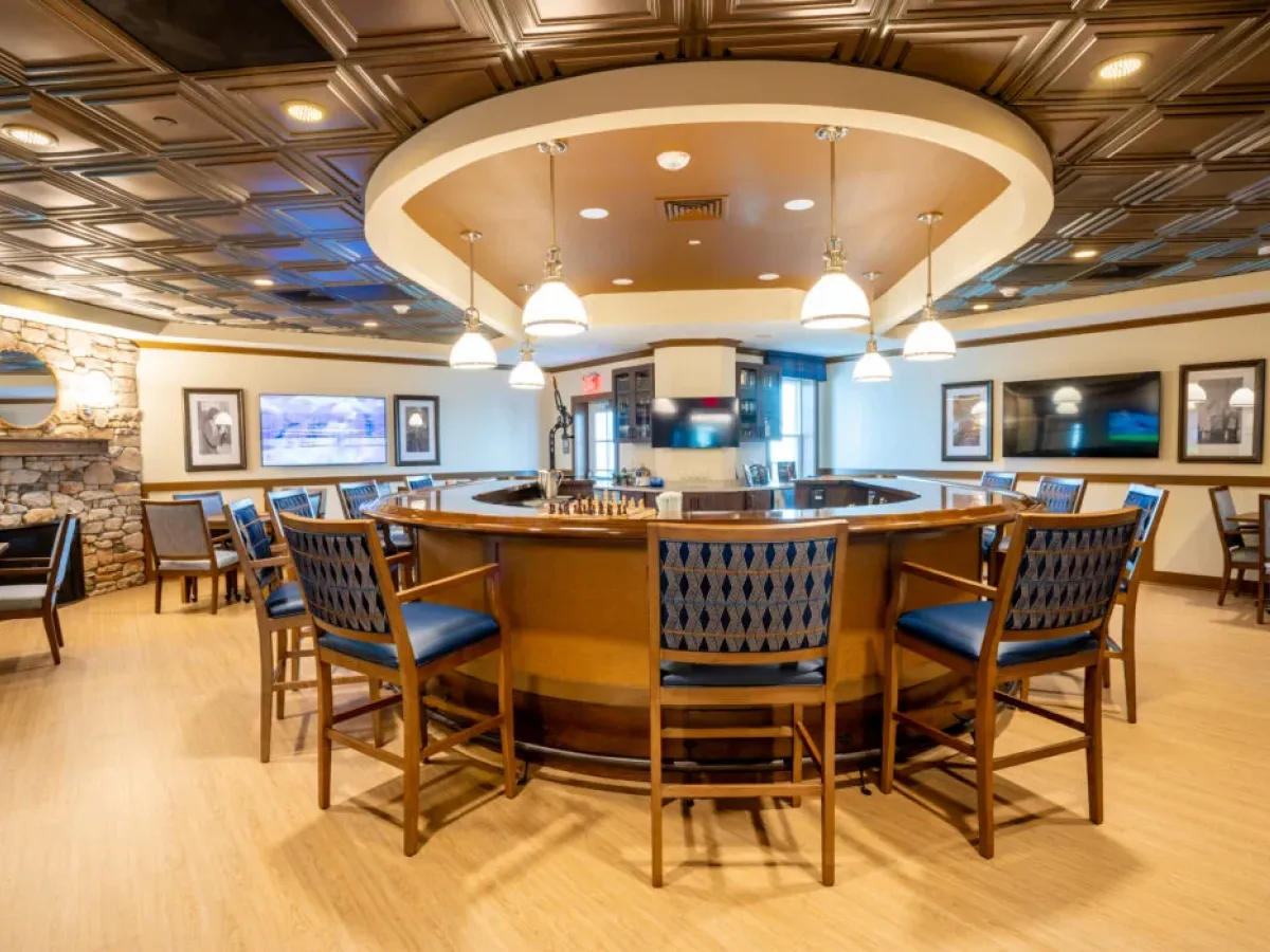 Westrum Corporate A round wooden bar in Azalea, with blue cushioned chairs, hanging lights, TVs, stone walls, and framed art in a modern lounge.