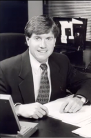 Westrum Corporate Smiling man in a suit sits at a desk with papers, a laptop, and office items in a professional office setting.