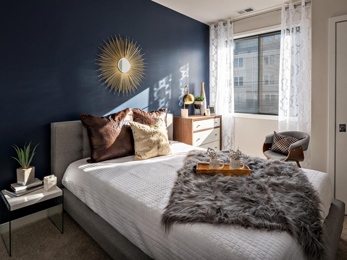 Westrum Corporate Modern bedroom at Pointe at 31 Apartments in Brewerytown featuring a navy accent wall, sunburst mirror, and cozy bed with fur throw.