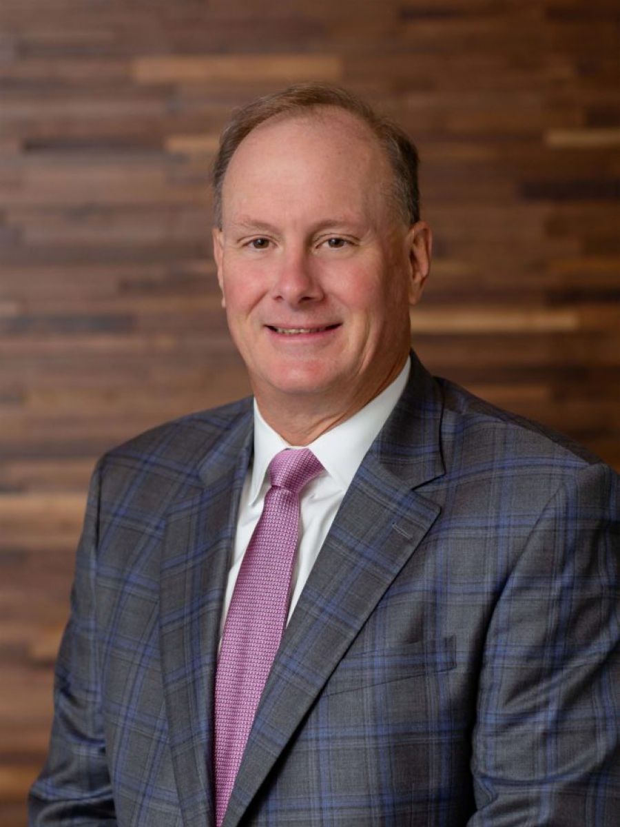 Westrum Corporate John Westrum, in a checked suit and pink tie, smiles while standing before a wooden wall background.
