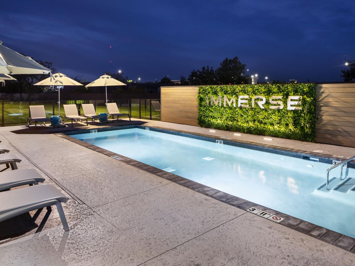 Westrum Corporate Modern outdoor pool at night at Luxor Wilmington, with white lounge chairs, umbrellas, and a glowing "IMMERSE" green wall sign.