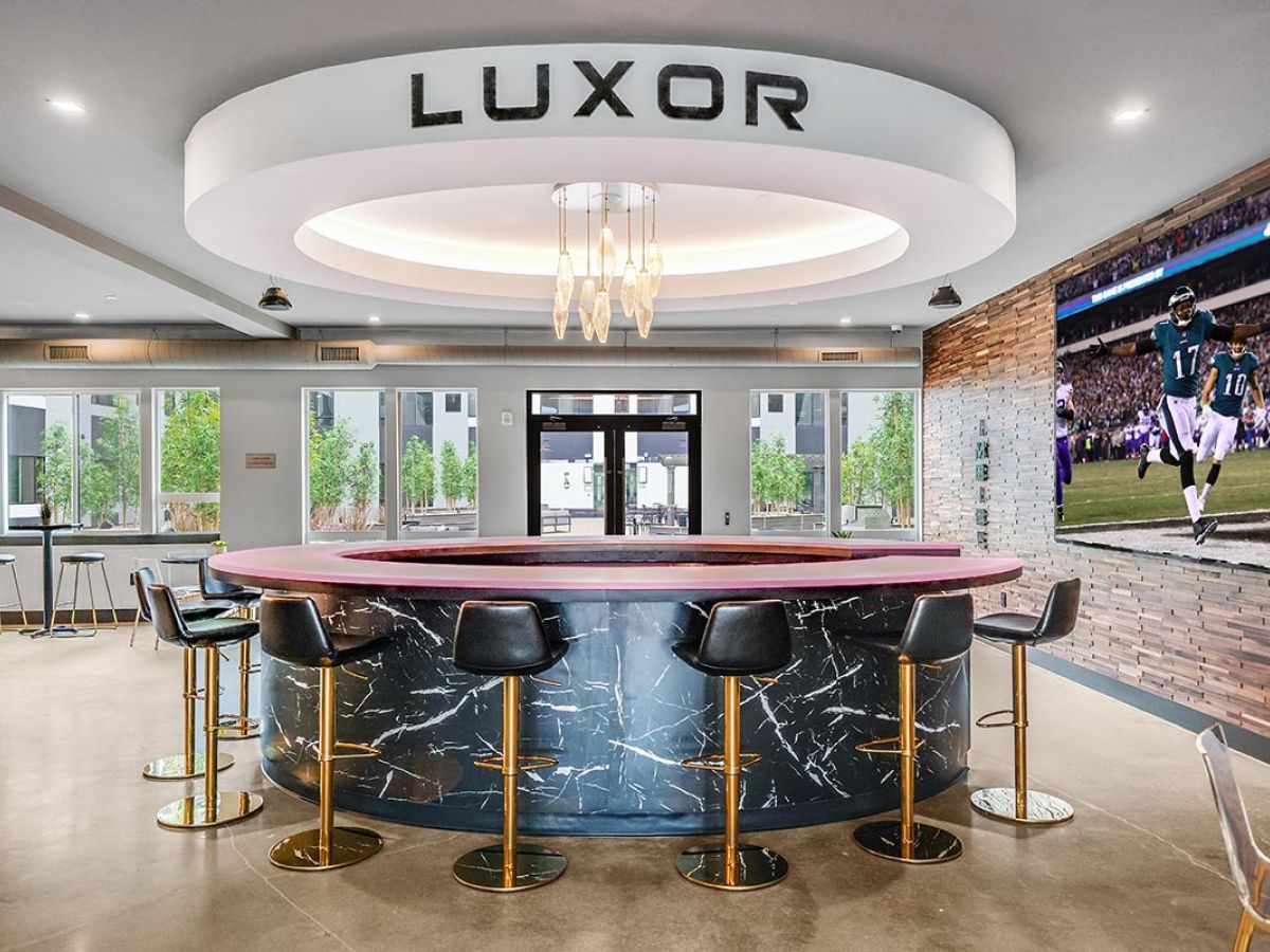 Westrum Corporate Modern lounge in West Chester with a circular black marble bar, “LUXOR” sign, barstools, and football on TV against a wood accent wall.
