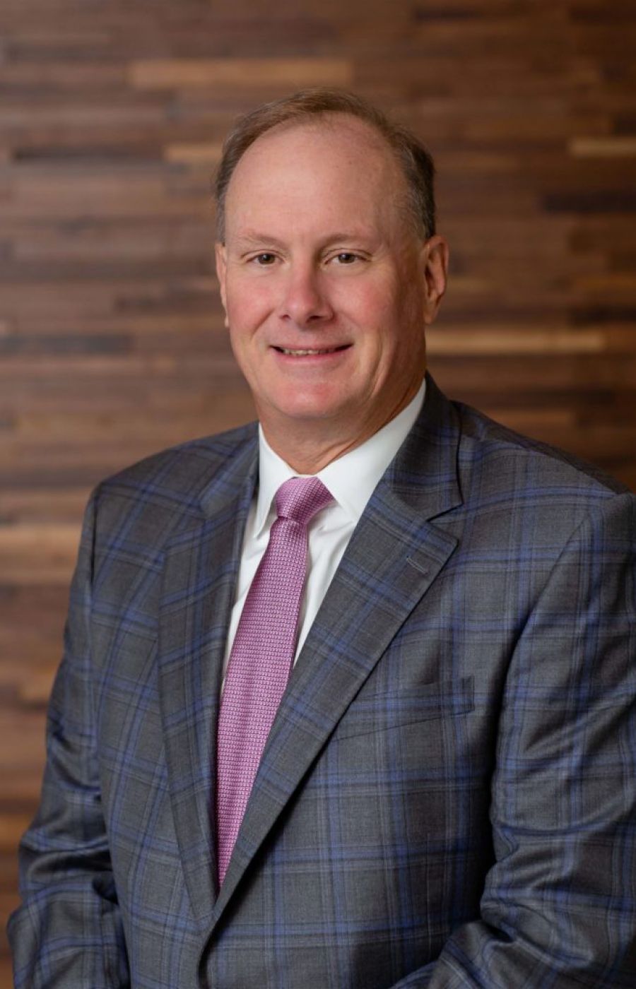 Westrum Corporate John Westrum, in a checked suit and pink tie, smiles while standing before a wooden wall background.