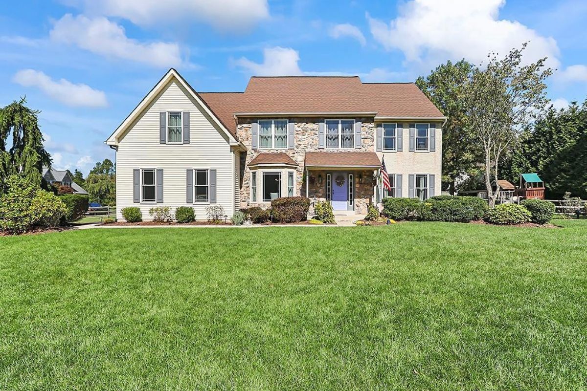 Westrum Corporate Charming two-story Buckingham Place home with stone and siding, lush green lawn, shrubs, and a bright sky dotted with clouds.