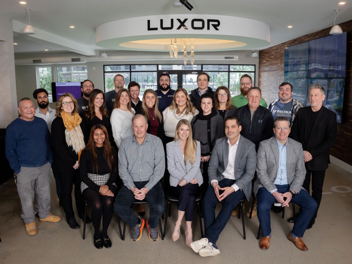 Westrum Corporate A group of 26 people posing and smiling together indoors under a sign that reads “LUXOR.”.