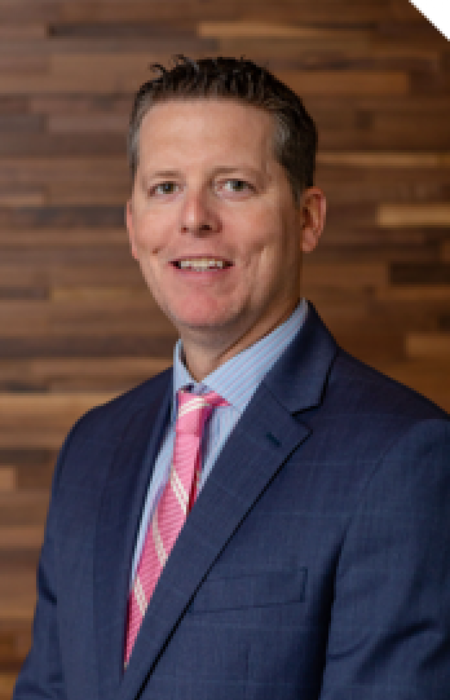 Westrum Corporate Brian McKenzie, in a blue suit and pink tie, stands smiling at the camera before a wooden wall.