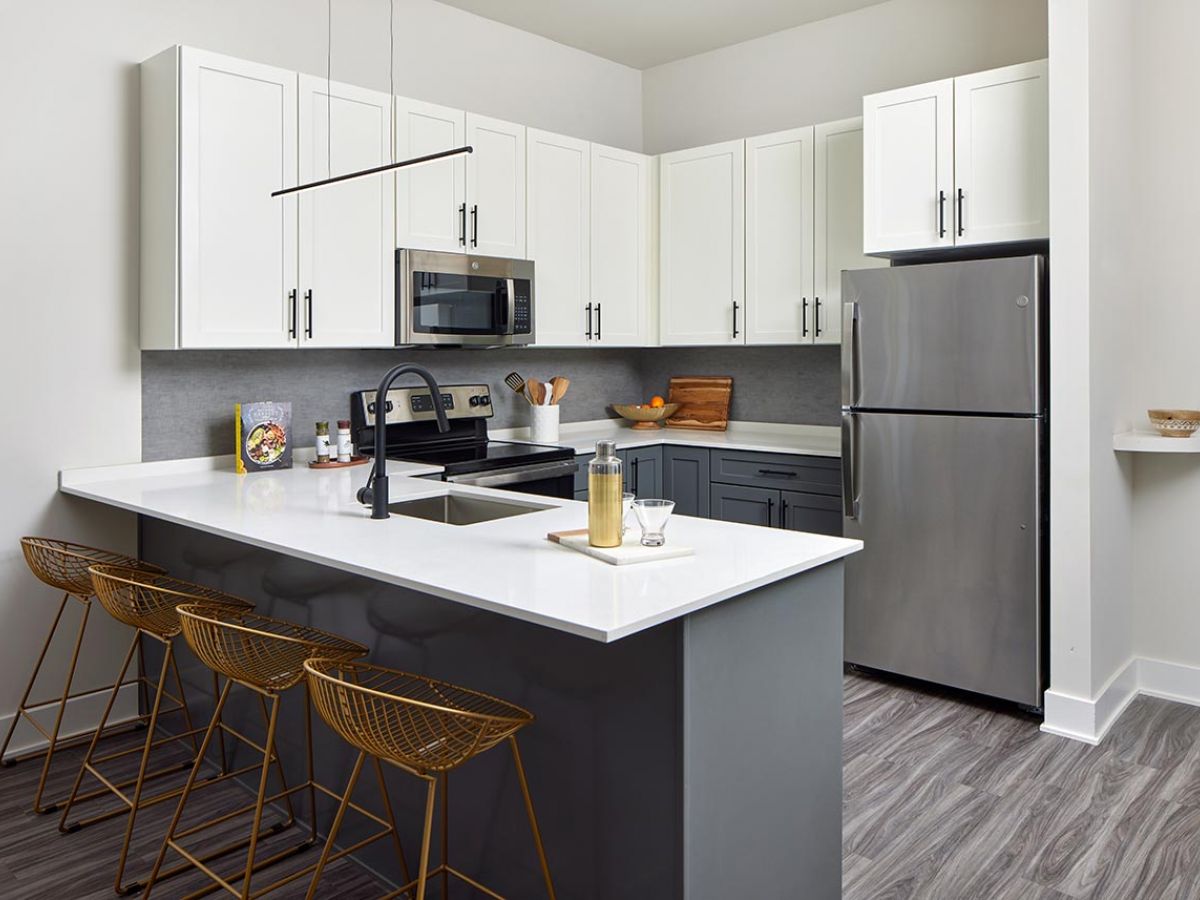 Westrum Corporate Modern kitchen at Luxor Wilmington boasts white cabinets, stainless steel appliances, and a sleek gray island with gold bar stools.