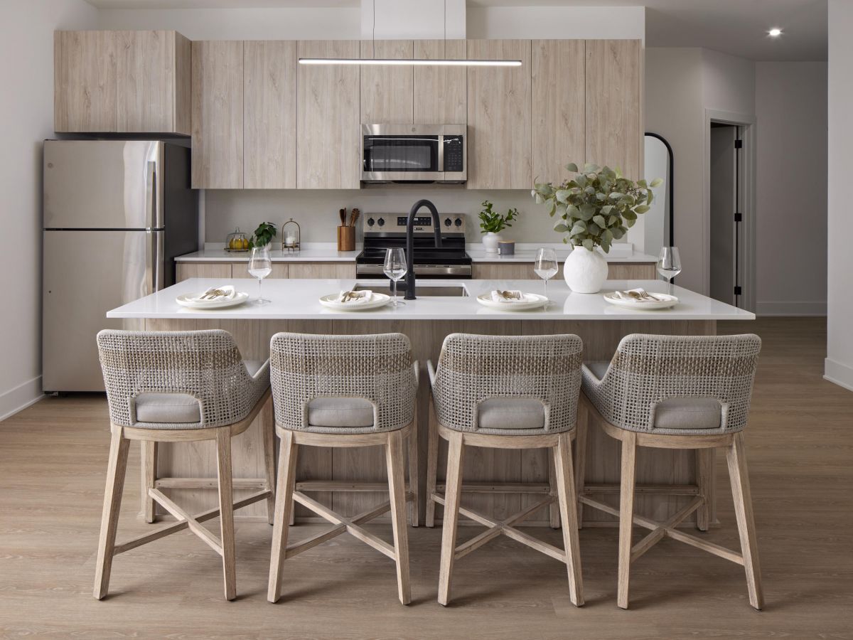 Westrum Corporate Modern kitchen at Foundry Flats features light wood cabinets, a white island, woven bar stools, and minimalist decor with greenery.
