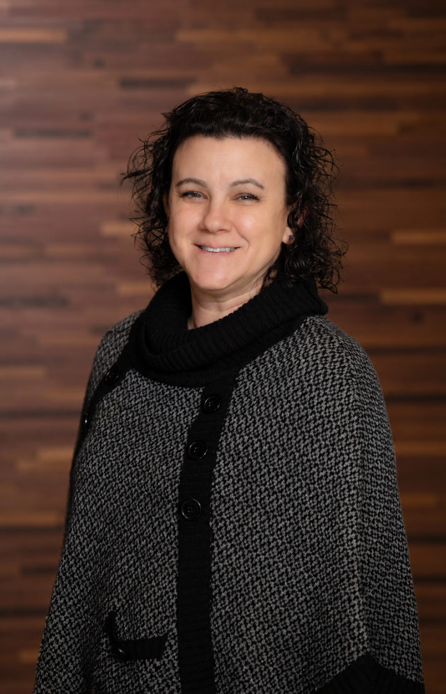 Westrum Corporate Christine Yordy, a woman with curly dark hair, stands smiling in a gray and black sweater before a wooden wall background.